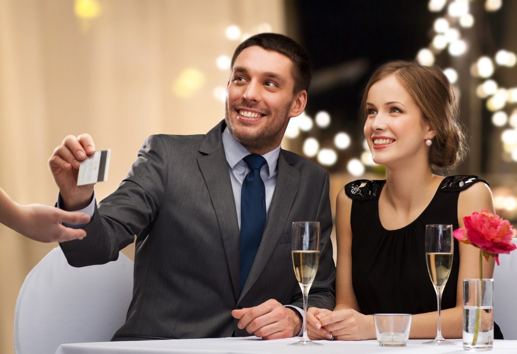 Man and woman happily paying for restaurant meal with a credit card, glasses of champagne and festive lights in the background.